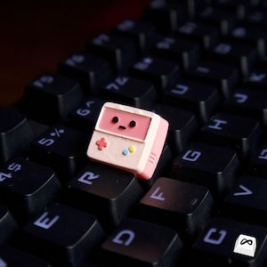 Könnte beinhalten: Eine rosa-weiße, individuelle Tastenkappe in Form einer Retro-Handheld-Spielkonsole mit kleinem Bildschirm, Steuerkreuz und farbigen Knöpfen. Die Tastenkappe befindet sich auf einer schwarzen Tastatur.