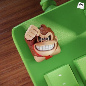 Könnte beinhalten: Nahaufnahme einer Donkey Kong-Themen-Tastenkappe. Die Tastenkappe zeigt einen braunen Affenkopf mit breitem Grinsen, roter Krawatte und geballter Faust. Die Tastenkappe befindet sich auf einer grünen Tastatur.