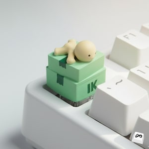May include: A light green artisan keycap featuring two stacked boxes with the letters "IK" and a small beige figure lying on top. The keycap is on a white keyboard.