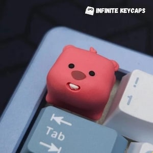 May include: A pink keycap shaped like a cartoon animal with a surprised expression. The keycap is on a keyboard with a blue and white color scheme. The keycap is labeled "Tab".