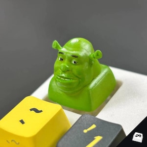 Puede incluir: Una tecla personalizada verde con la temática de Shrek, con la cara del personaje, colocada en un teclado. Está rodeada de otras teclas de colores, incluyendo amarillas y grises. El diseño es detallado y caprichoso.