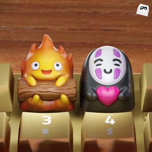 Puede incluir: Dos teclas personalizadas con personajes animados. Una es una criatura naranja llameante que sostiene un tronco, y la otra es una figura negra con una cara blanca y un corazón rosa. Las teclas están en un teclado dorado.