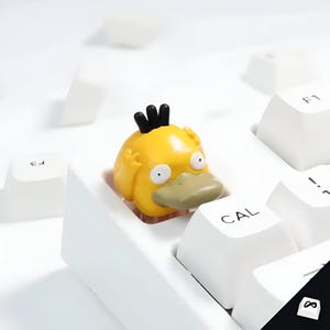 Puede incluir: Una tecla amarilla de Psyduck con pelo negro y pico verde claro se encuentra en un teclado blanco. La tecla es un diseño personalizado, que añade un toque único a una configuración informática. Las teclas del teclado son blancas con letras negras.