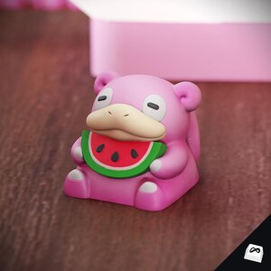 May include: A pink Slowpoke figurine holding a watermelon slice. The figurine has a light tan snout, black eyes, and small ears. The watermelon slice is green with red and black details.