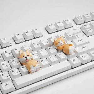 May include: A white mechanical keyboard with custom keycaps shaped like a corgi and a cat. The corgi keycap is winking, and the cat keycap is orange and white. The keyboard has white keycaps with black lettering.