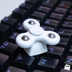 May include: A white fidget spinner with three rounded arms and silver ball bearings, resting on a black computer keyboard. The keys are dark with white lettering.