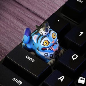 May include: A blue and black keyboard keycap shaped like a cartoon tiger with a small creature on its head. The tiger has yellow eyes, a pink tongue, and black stripes. The keycap is on a black keyboard.