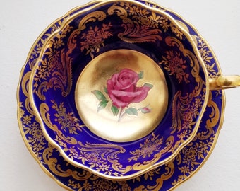 May include: A cobalt blue teacup and saucer set with gold filigree and a gold interior. The teacup features a painted pink rose in the center. The set has a scalloped edge and is likely vintage or antique.