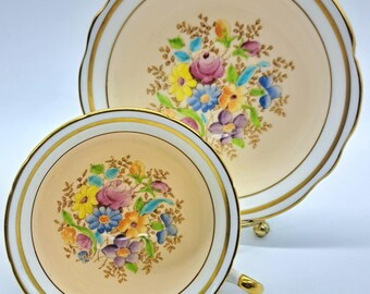 May include: A vintage teacup and saucer set. The set features a cream-colored background with a floral design in shades of pink, yellow, blue, and purple. Gold trim accents the edges of the cup and saucer, and the cup has a gold handle.