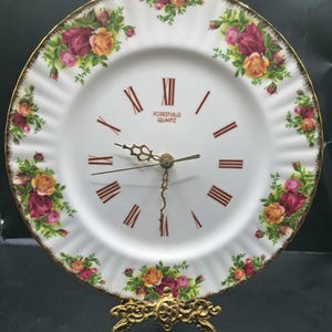 Puede incluir: Un reloj de plato blanco con un diseño floral de rosas en tonos rosa, amarillo y verde. La esfera del reloj presenta números romanos y el texto "FORESTVILLE QUARTZ". El reloj está sostenido por un soporte de metal dorado.