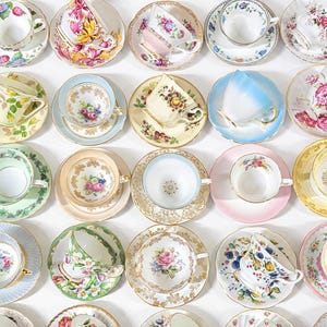 May include: Overhead shot of a collection of vintage teacups and saucers. Each cup and saucer features unique floral patterns in various colors, including pink, blue, yellow, and green. The teacups are arranged on a white surface.