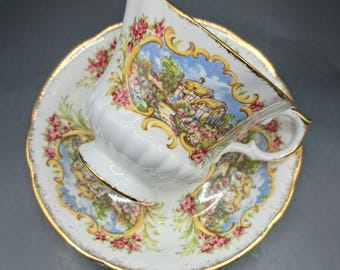 May include: White teacup and saucer set with a gold trim. The cup and saucer feature a scenic landscape design with pink flowers and a gold border. The cup has a curved handle and a scalloped edge. The saucer has a matching design.