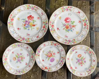 May include: A collection of five white porcelain plates with floral patterns. Each plate has a gold rim and a decorative border of flowers and leaves. The centre of each plate displays a bouquet of colourful flowers, including red, pink, yellow, and blue.