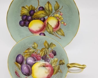 May include: A vintage teacup and saucer set with a delicate blue and white floral design. The set features a hand-painted design of fruit, including plums, peaches, and blackberries.