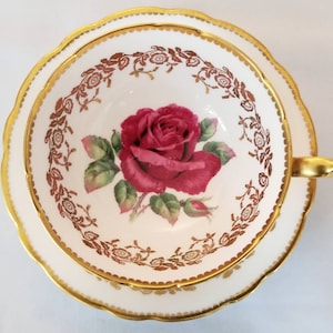 Może przedstawiać: Biały kubek do herbaty i spodek z pozłacanym rantem i wzorem czerwonej róży. Różę otacza złoty wzór kwiatowy.