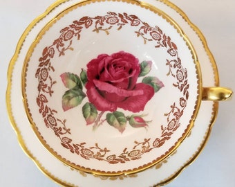 May include: A white teacup and saucer with a gold rim and a red rose design. The rose is surrounded by a gold floral pattern.