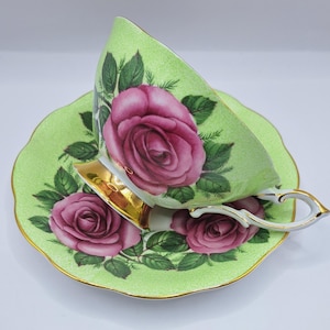 Może przedstawiać: Zestaw filiżanki i spodka do herbaty w stylu vintage. Filiżanka i spodek są jasnozielone z kwiatowym wzorem róż w odcieniach różu i zieleni. Filiżanka ma złoty uchwyt i obramowanie.