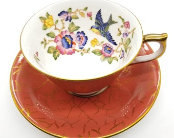 May include: A white teacup and saucer with a floral and bird design. The cup has a gold rim and the saucer has a coral red base with gold accents.