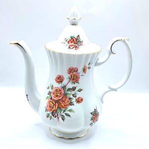 Puede incluir: Cafetera de porcelana blanca con tapa, adornada con un motivo floral de rosas rosas, naranjas y verdes. La cafetera presenta un pico curvo, un asa y ribetes dorados. Una pieza clásica para servir café o té.