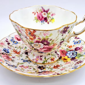 Könnte beinhalten: Ein Vintage-Teetassen- und Untertassen-Set mit floralem Muster auf weißem Hintergrund und einem zarten Blumenmuster in Rosa-, Lila-, Blau- und Gelbtönen. Tasse und Untertasse haben einen goldenen Rand.