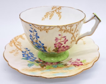 Può includere: Un set vintage di tazza da tè e piattino con un motivo floreale. La tazza ha una base verde e il piattino ha un bordo ondulato. Il motivo floreale presenta fiori blu e rosa su uno sfondo crema.