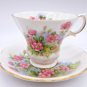 Peut inclure: Une tasse à thé et une soucoupe blanches avec un motif floral. La tasse et la soucoupe sont décorées de roses roses et de feuilles vertes sur un fond bleu clair. Les bords de la tasse et de la soucoupe sont bordés d'or.