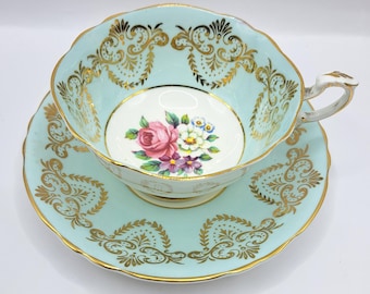 May include: A vintage teacup and saucer set. The cup and saucer are a light blue with gold filigree and trim. The center of the cup features a floral design with a pink rose and other colorful flowers. A classic tea set.