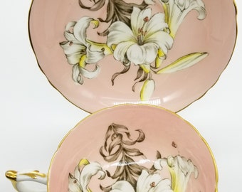 May include: A pink teacup and saucer set featuring a white floral design. The flowers are lilies with brown stems and yellow accents. The set has a gold trim.
