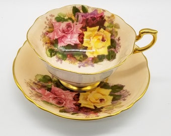May include: A vintage teacup and saucer set. The cup and saucer are a pale peach color with a gold trim. The cup and saucer are decorated with a floral pattern of pink, yellow, and purple roses. The handle of the cup is gold.