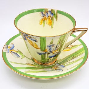 May include: A vintage teacup and saucer set. The cup and saucer are cream-coloured with a green border and gold trim. The set is decorated with blue and yellow iris flowers and green leaves.