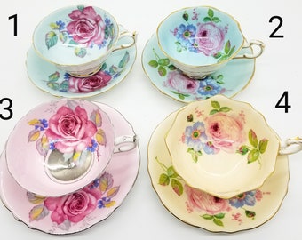 May include: Four teacups and saucers with floral patterns. The cups are in different colors: light blue, pink, and cream. Each cup has a different floral design featuring roses and other flowers.