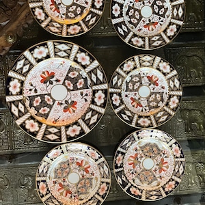 May include: Six decorative plates with a floral design. The plates have a white centre with orange and red flowers, surrounded by a black and white geometric pattern and a gold rim.