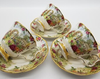 May include: Three white teacups and saucers with a floral pattern and gold trim. The cups and saucers feature a cottage scene with a garden and flowers.