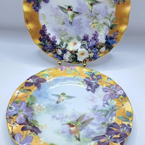 May include: Two decorative plates with gold rims and scalloped edges. Each plate features a painted scene of hummingbirds, white roses, and purple flowers. The plates are displayed on gold stands against a white background.