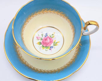 Può includere: Un set vintage di tazza da tè e piattino con un design floreale blu e bianco. La tazza ha un bordo dorato e un delicato motivo floreale al centro. Il piattino ha un design blu e bianco abbinato con un bordo dorato.