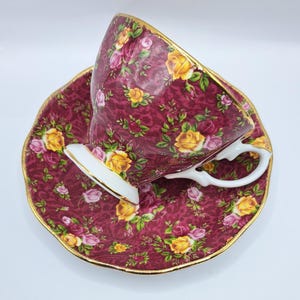 Puede incluir: Un juego de taza y platillo de color burdeos con un estampado floral de rosas amarillas y rosas. La taza y el platillo tienen ribetes dorados y un asa blanca. El juego está diseñado para el té.