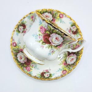 Può includere: Servizio da tè bianco con tazza e piattino con un motivo floreale a rose rosa, rosse e bianche. I bordi della tazza e del piattino sono rifiniti in oro. La tazza da tè è appoggiata sul piattino.