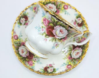 May include: White teacup and saucer set with a floral rose pattern in pink, red, and white. The edges of the cup and saucer are trimmed with gold. The teacup is resting on the saucer.