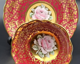 May include: A red and gold teacup and saucer set with a pink rose design. The saucer has a scalloped edge and the cup has a handle.