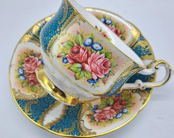 May include: A vintage teacup and saucer set. The teacup and saucer are decorated with pink roses, blue accents, and gold trim. The teacup has a curved handle and sits on a matching saucer with a scalloped edge. The set has a teal and gold design.