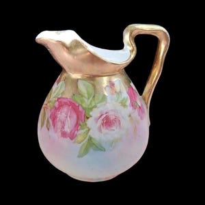 Puede incluir: Una jarrita de porcelana con un diseño floral pintado a mano de rosas rosas y blancas con hojas verdes. La jarra tiene un borde y un asa pintados en oro, con un cuerpo redondeado que pasa de una base blanca a una sección superior dorada.
