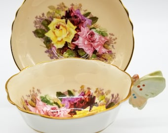 Può includere: Un set da tè bianco con una tazza e un piattino con un motivo floreale con rose rosa, gialle e viola. La tazza da tè ha un manico a forma di farfalla.