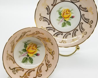 May include: A pink teacup and saucer with a gold trim and a yellow rose design. The saucer has a scalloped edge.