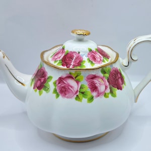 Pode incluir: Bule de cerâmica branca com tampa, adornado com padrões florais de rosas rosa e vermelhas e detalhes dourados. O bule tem um bico e uma alça curvos. Uma peça decorativa para a cozinha.