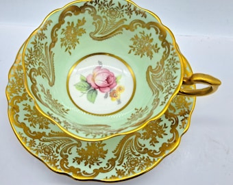 Peut inclure: Une tasse à thé et une soucoupe vintage avec un délicat motif floral vert et or. La tasse présente une seule rose rose avec des feuilles vertes et un bord doré. La soucoupe a un motif floral assorti avec un bord doré.