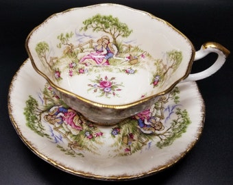 May include: Cream-colored teacup and saucer set with a romantic pastoral scene. The cup and saucer feature a gold trim and a detailed illustration of a couple in a field. The set is decorated with pink, blue, and green floral accents.