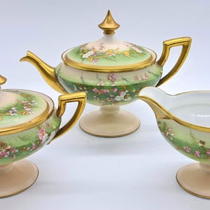 May include: A three-piece porcelain tea set comprising a teapot, sugar bowl, and creamer. The set has a cream base, gold accents, and a green and floral design. The teapot has a gold spout and handle.