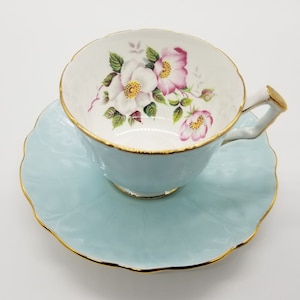 May include: A light blue teacup and saucer set with a floral design. The cup and saucer have a gold trim. The floral design features pink and white roses.