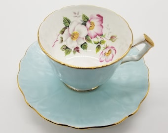 Può includere: Un set da tè composto da una tazza e un piattino di colore azzurro chiaro con un motivo floreale. La tazza e il piattino hanno un bordo dorato. Il motivo floreale presenta rose rosa e bianche.