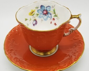 May include: A vintage teacup and saucer set in a bright orange color with a gold trim. The teacup features a delicate floral design with a blue flower and yellow accents.